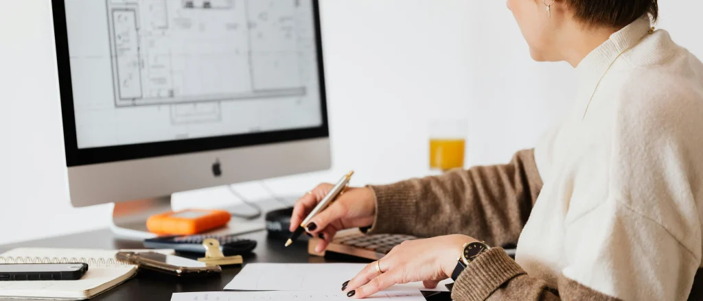 Evita errores en planos de casas revisando tus diseños en papel y pantalla Arquitecta revisando planos de casas en papel con diseño técnico en pantalla